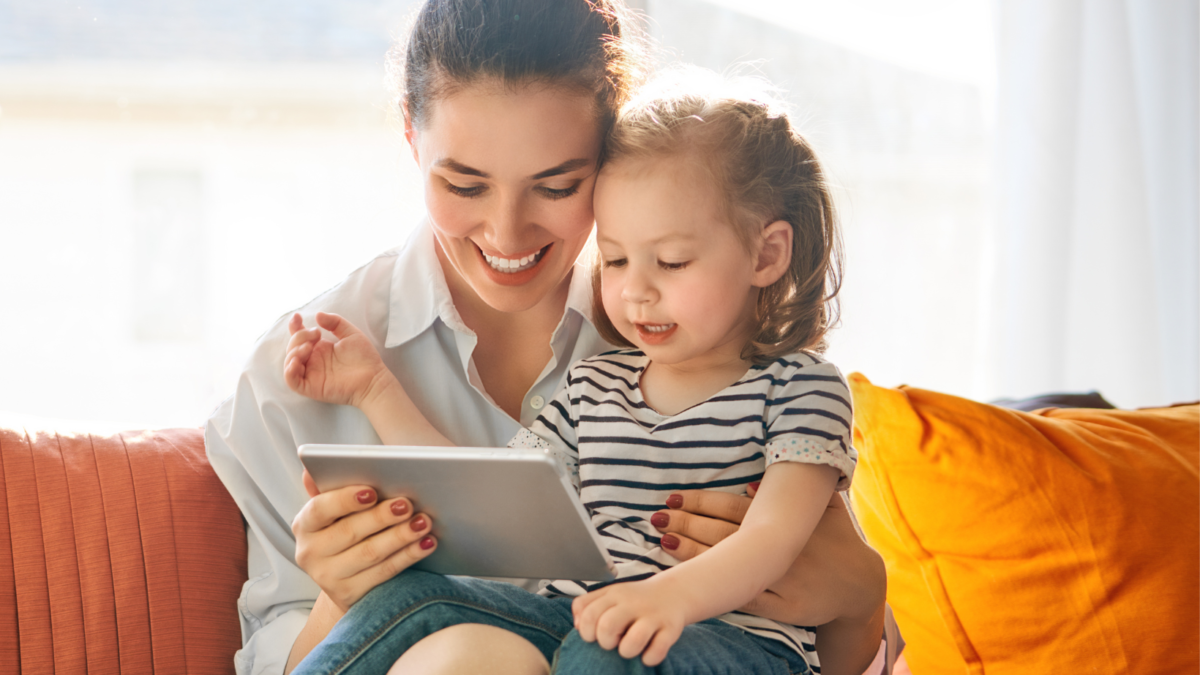 Mamma e bambina sorridenti guardano insieme un tablet sedute sul divano