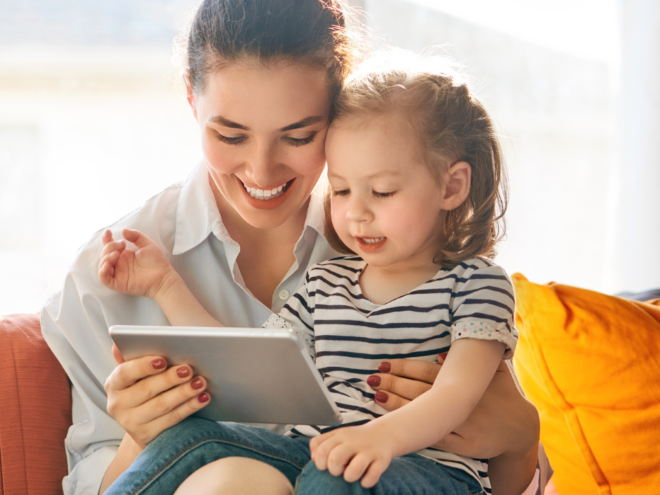 Mamma e bambina sorridenti guardano insieme un tablet sedute sul divano
