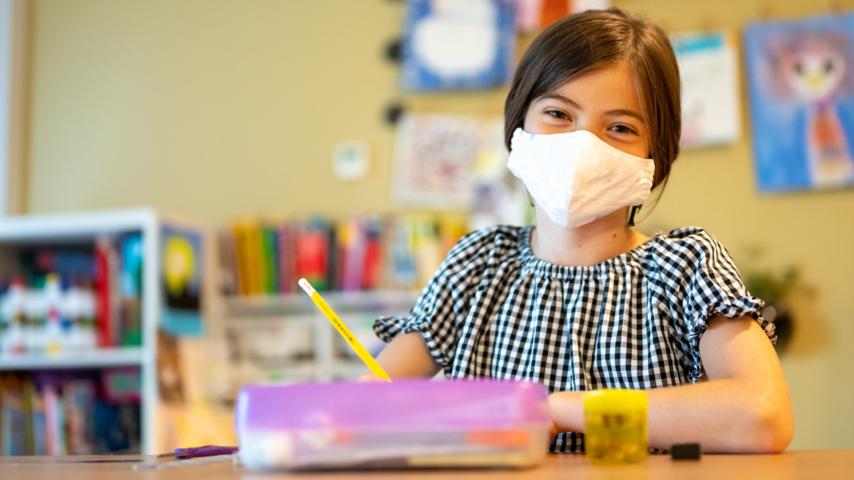 Bambina con mascherina seduta al banco di scuola, con disegni colorati alle pareti