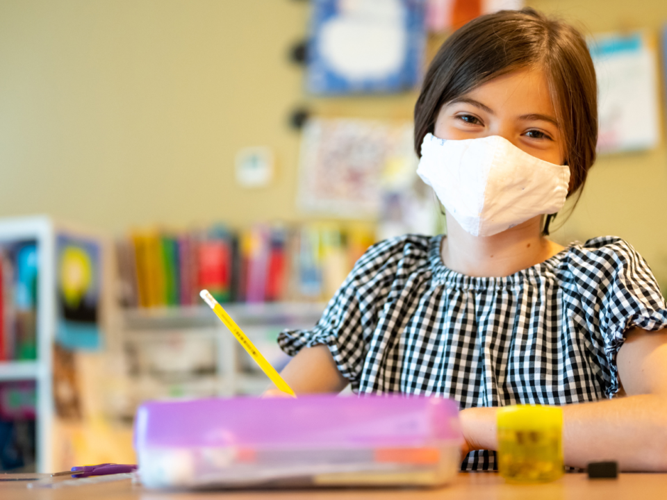 Bambina con mascherina seduta al banco di scuola, con disegni colorati alle pareti