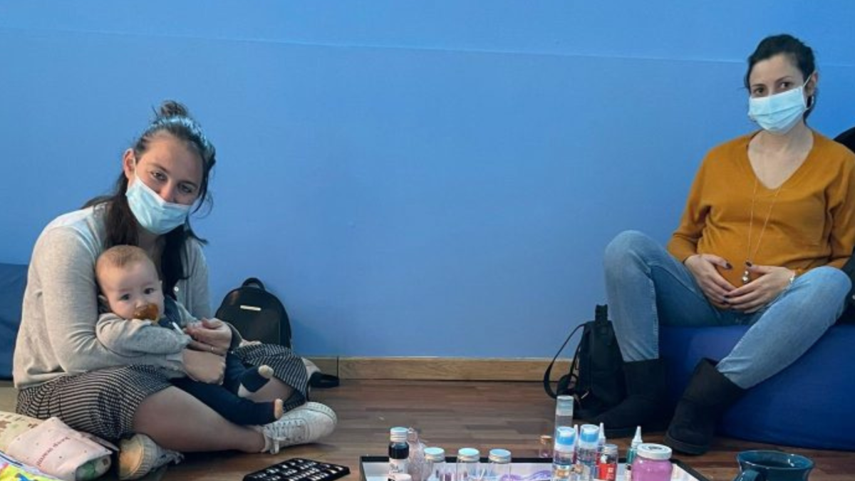 Mamme con mascherina sedute a terra durante un incontro del gruppo neomamme, una con un bebè in braccio