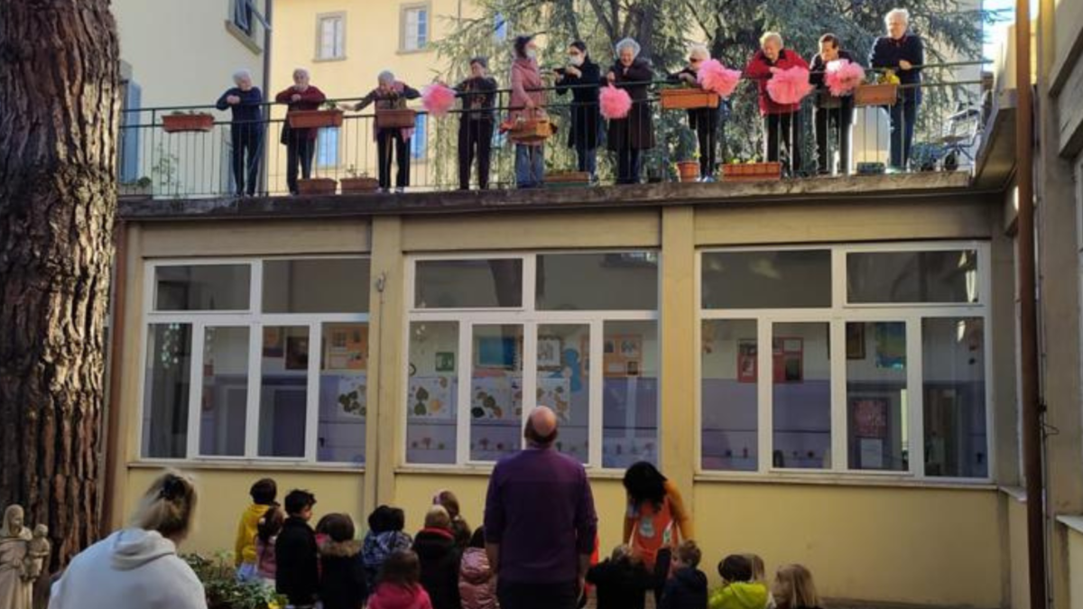 Anziani affacciati al balcone salutano i bambini della scuola riuniti nel cortile