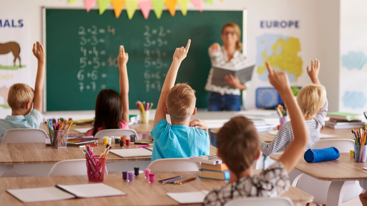 Alunni in classe alzano la mano per rispondere durante una lezione con l'insegnante alla lavagna