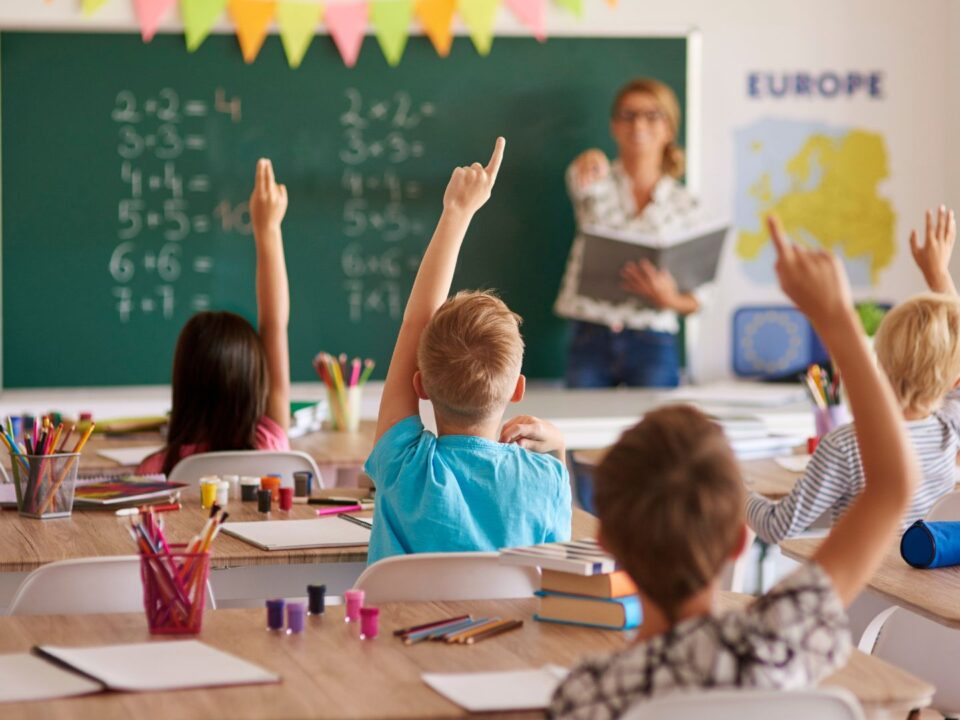 Alunni in classe alzano la mano per rispondere durante una lezione con l'insegnante alla lavagna