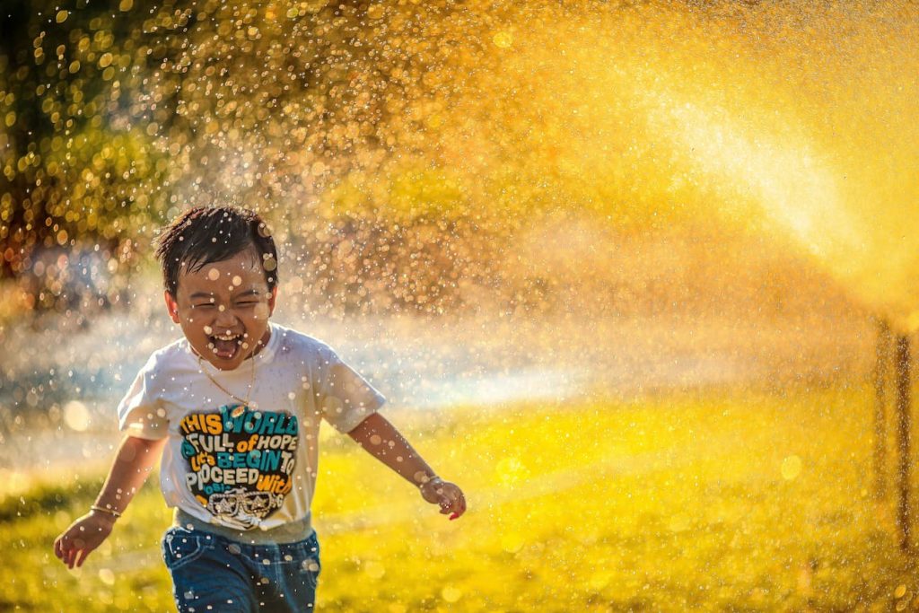 Bambino sorridente corre bagnato tra gli spruzzi d'acqua di un irrigatore in un giorno d'estate